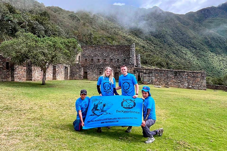 Group in blue shirts poses on grass holding a TreXperience banner in front of stone walls and mountains, Choquequirao Trek 5 Days 4 Nights | TreXperience