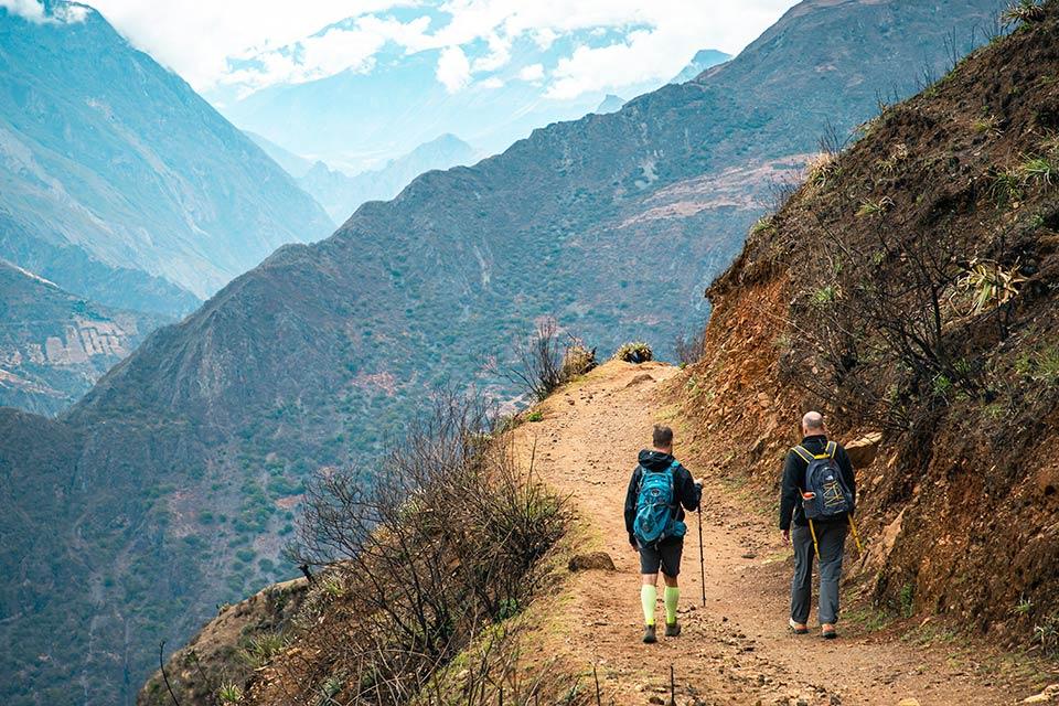 Two hikers walk along a dirt trail on a hillside, with a deep valley and mountains in the background, Choquequirao Trek 5 Days 4 Nights | TreXperience