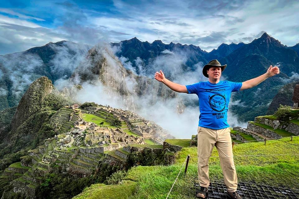 Turista con brazos abiertos en mirador con Machu Picchu y montañas entre nubes al fondo, Huchuy Qosqo y Camino Inca Corto 4 días | TreXperience