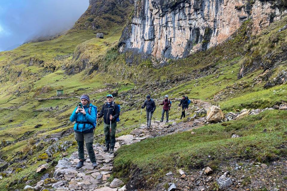 Group of hikers with backpacks and trekking poles walking along a mountain trail beside a rocky cliff during a hike | TreXperience