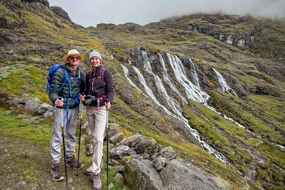 Two hikers with backpacks and trekking poles posing in front of a waterfall in a mountain landscape during a hike | TreXperience