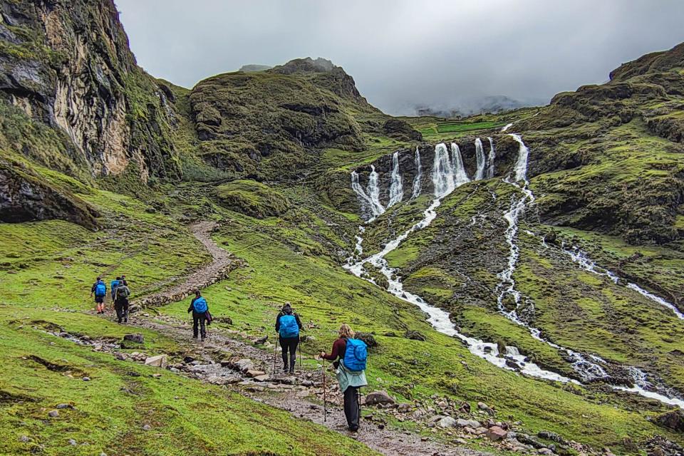 Group of hikers with backpacks walking toward a waterfall along a mountain trail during a hike | TreXperience