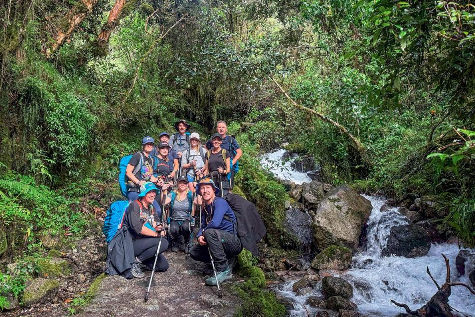 Group of hikers with backpacks and trekking poles posing beside a stream on a trail surrounded by Andean forest during a hike | TreXperience