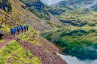 Caminantes con mochilas avanzan por un sendero junto a una laguna verde en valle de montaña, Trek Lares a Machu Picchu 4 días | TreXperience