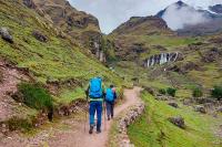 Dos caminantes avanzan por un sendero en valle verde con cascadas al fondo y nubes bajas, Trek Lares a Machu Picchu 4 días | TreXperience