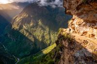Narrow cliffside trail carved into rock above a deep valley, with a hiker and mountains ahead, Choquequirao Machu Picchu 7 Days Trek | TreXperience