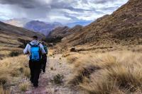 Dos caminantes avanzan por un sendero de montaña entre ichu y laderas, con montañas al fondo, Huchuy Qosqo y Camino Inca Corto 4 días | TreXperience