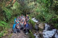 Group of hikers with backpacks and trekking poles posing beside a stream on a trail surrounded by Andean forest during a hike | TreXperience